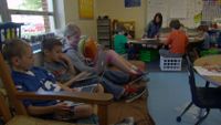 Students in a classroom with teacher working with another group of students in the corner.