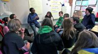 A student is speaking in front of the classroom among his friends listening to him.