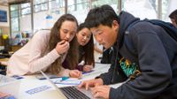 Three middle school students lean over a laptop while working on a class project.