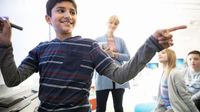 Smiling boy at whiteboard pointing to his classmates while his teacher watches.