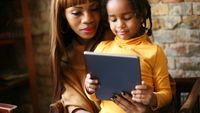 A daughter sitting on her mother's lap, both looking at a tablet that the daughter is holding