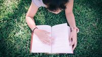 A middle school girl reading a book in a park on the grass