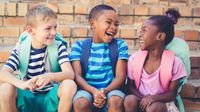 Three elementary school children wearing backpacks, sitting on the school steps, talking and laughing together