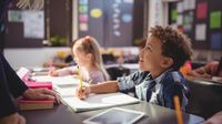 A young boy sitting at his desk, looking up at his teacher who is talking to him, and smiling