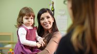 A mother smiling and holding her young daughter in her arms while talking to her daughter's teacher