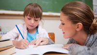 Teacher observing a young student as she writes in a notebook in class