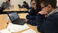 Two students looking at data on a laptop with worksheets scattered in front of them