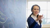 Teacher standing next to a blackboard that has a diagram drawn on it. She is pointing off-camera.