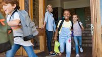 A teacher letting her students out the front door of the school on the last day of the school year. The kids are running with smiles on their faces, and the teacher is smiling as she watches them go.