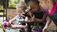 Three kindergarten students doing a science lab. The girl in the middle is looking into a microscope and the other two girls are looking on and writing things down on paper with markers.