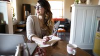 A woman sitting at her kitchen table with a cup of tea, writing in a notebook and looking thoughtfully out the window