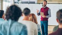 A teacher gesturing in front of a whiteboard to a room of his fellow teachers and faculty