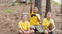 Three elementary school children kneeling on the ground in a forest. One is holding a pair of binoculars and pointing upward toward the trees. The other two are looking up to where he's pointing.