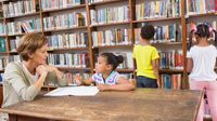 A librarian talking to a young girl at a table while two students browse the stacks behind them