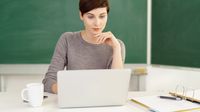 A teacher using her laptop in a classroom. She's sitting in front of a blackboard and has a notebook next to her.