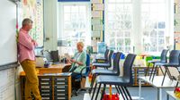 A teacher and a principal meeting in a classroom after school. The teacher is sitting at her desk and the principal is standing, leaning against the whiteboard.