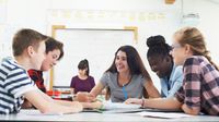 A group of five students working together in a classroom, their teacher sitting at a desk doing paperwork in the background