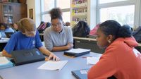 Three high school students at table looking at spiral-bound notebook.