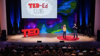 A male and female adult are standing on a stage with a large audience in front of them. Standing on the stage are three big, red letters spelling, "TED." On a screen behind them says, "Ted-ed clubs."