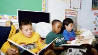 Three young boys are sitting on a couch in their classroom, reading.