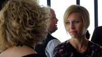 Two women are standing in a room, talking to each other. Other adults and standing and talking in the background.