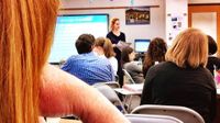 High school students are sitting at desks in foldable chairs. A teacher is standing at the front of the class next to a large screen on the white board. The screen is displaying a presentation slide that says, "Language Acquisition."