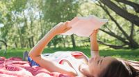 A young girl is lying on her back with a book in her hands. She's lying on a red and white checkered blanket on the grass. She's surrounding by grass, trees, and the sun.