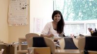 A teacher is sitting by herself in her classroom by a window. At her desk, with a pencil in hand and paper in front of her, she's smiling at the camera.