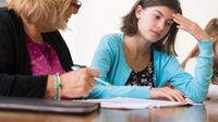 A short-haired, blond female teacher in a pink shirt and black blouse is sitting next to a brunette teenager girl, helping her study.