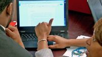 Teacher crouching down at a desk helping a student with his composition on a computer
