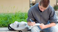 Older boy sitting on a large cement planter in a school courtyard writing in a spiral notebook