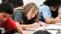 Students are hunched over their desks, writing in notebooks, with their teacher looking upon them.