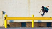 A young teenage boy in a teal t-shirt and brown pants is in the air on a skateboard. Both of his arms are extended out to his sides and his knees are up to his chest. He's hovering over a yellow beam against an off-white brick wall.