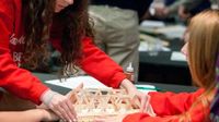 Two students are sitting, and one student is standing, all their hands are touching a popsicle stick bridge that they're gluing together.
