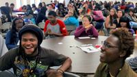 A large number of students are sitting at circular tables, filling the classroom.