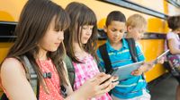Five young students are leaning against a bus all looking at mobile devices.
