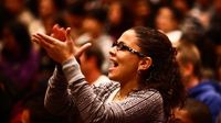 An adult woman is standing and clapping among a crowd of people sitting.
