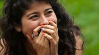 A closeup of a teenage girl with long, brown, curly hair, smiling, her hands covering her mouth.