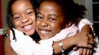 Two young girls are smiling and hugging each other, looking at the camera.