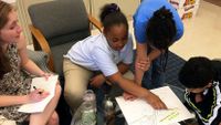 Two young girls and a boy are sitting around a circular glass table with their teacher, looking at a map.