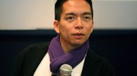 An adult male is sitting, holding a microphone, and speaking to someone. He's wearing glasses perched on top of his head, a white top, black coat, purple scarf, and a conference name tag on his chest.