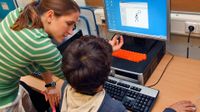 A teaching is standing next to a sitting student, talking to him and pointing at the desktop computer screen in front of him.