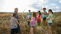 Nine students are standing in a vast field with a male teacher among tall, dry grass. They're placing hand-made windmills in the field.