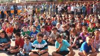 A lot of young students are sitting outside on a blacktop, with teachers standing behind them, all facing the same direction.
