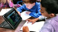 A group of elementary students are sitting at circular tables, sharing a tablet, and working on a paper assignment.