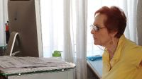 An older, red-headed woman with glasses and a yellow top is sitting in an office, looking at a computer screen.