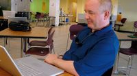A teacher is sitting at a small table, smiling at his laptop. His cell phone is beside him on the table, connected to his laptop.
