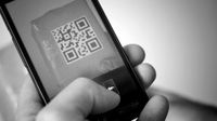 A black and white closeup of a hand holding a cell phone with a QR code on the screen.