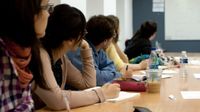 Six young adults are sitting on one side of a conference table looking towards the front of the room where a man is pointing at a whiteboard.