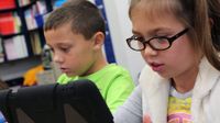 A young girl wearing glasses, pigtails, and a white vest over a gray, long sleeve shirt is sitting, looking at a tablet that's propped up on the table. She is sitting beside a boy in a bright green shirt who's also looking at a tablet.